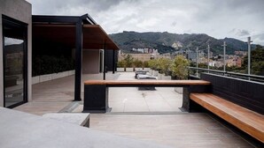 Une terrasse extérieure avec un banc en bois, un espace salon couvert et une vue sur le paysage urbain et les montagnes en arrière-plan.