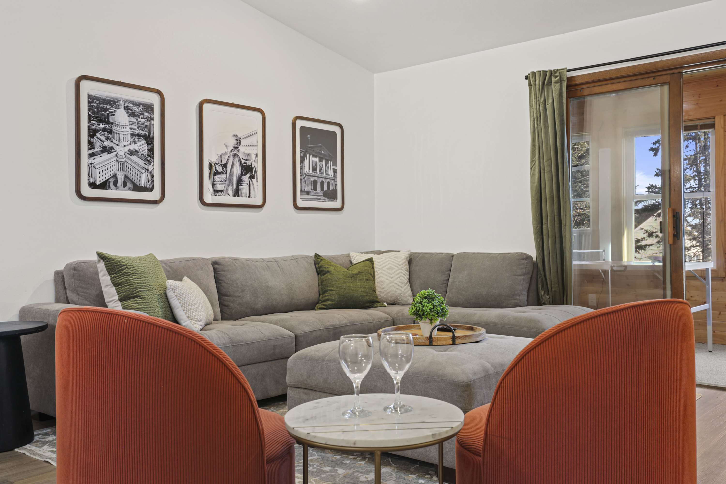 A living room with a grey sofa, two orange chairs, a small round table with two wine glasses, and three framed black-and-white photographs on the wall.