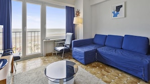A modern hotel room with a large window, a blue sofa, a round glass table, and a desk with a chair. - Apartment in Blankenberge With Sea Views (Blankenberge)