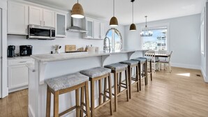 A modern kitchen with white cabinets, a stainless steel microwave, and a coffee maker. There are bar stools with cushioned seats and pendant lights above the island. - Experience luxury at Salt and Light in Ocean Isle Beach, perfect for 24 guests! (Ocean Isle)