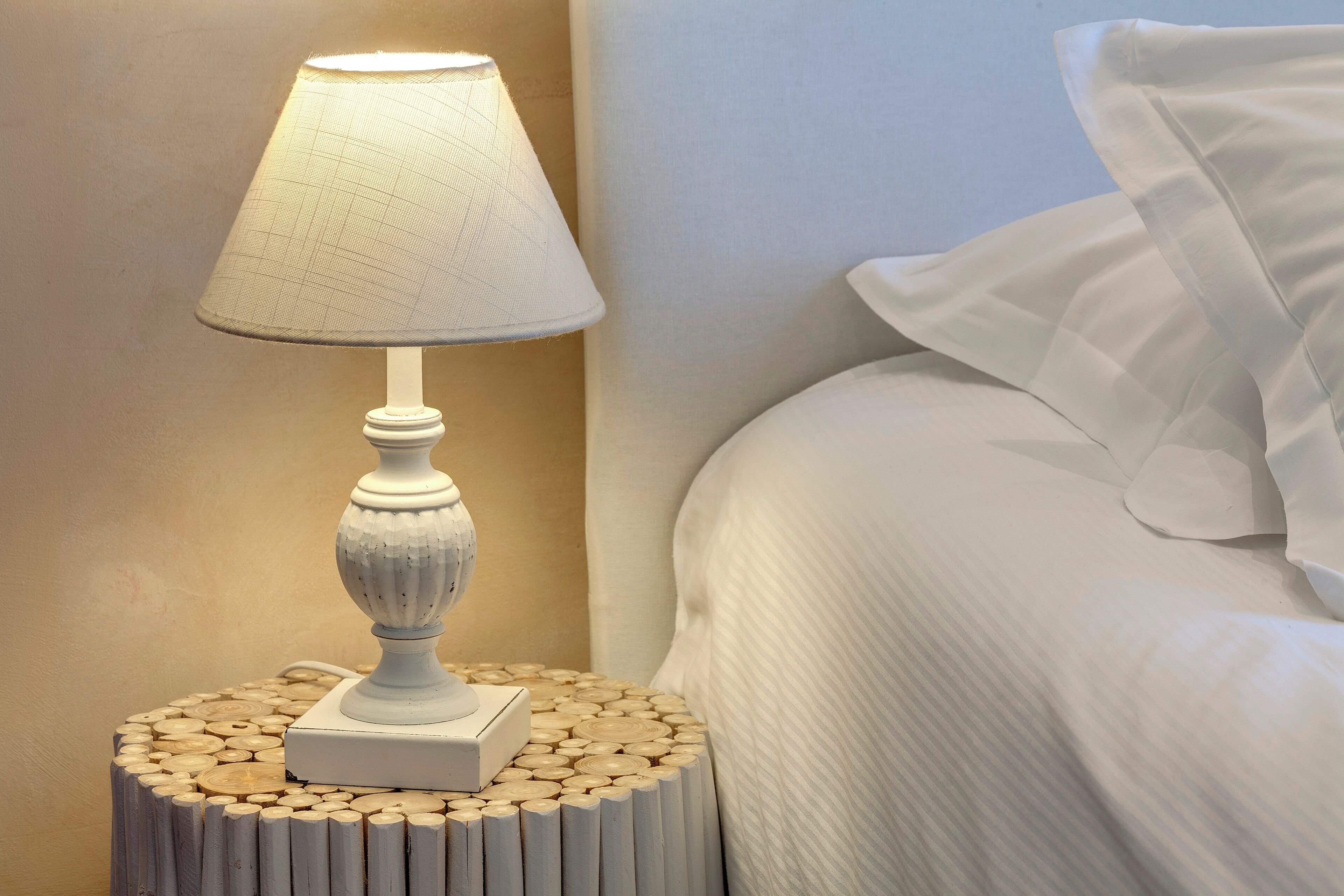 A bedside table with a lamp, a bed with white bedding, and a textured wall.