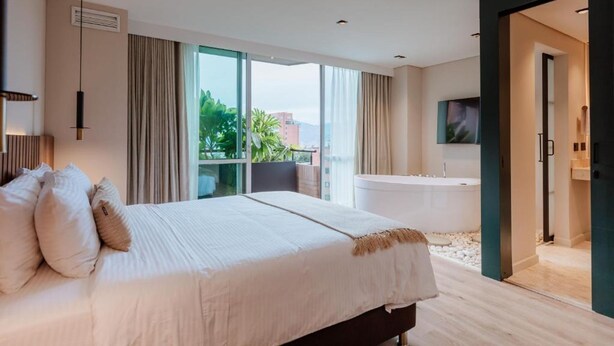 A modern hotel room with a large bed, a bathtub, and a view of greenery outside. - w Loft Estandar (Medellín)