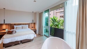 A modern hotel room with a large bed, a sitting area with a wooden headboard, a large window with a view of greenery, and a white bathtub. - w Loft Estandar Plus (Medellín)