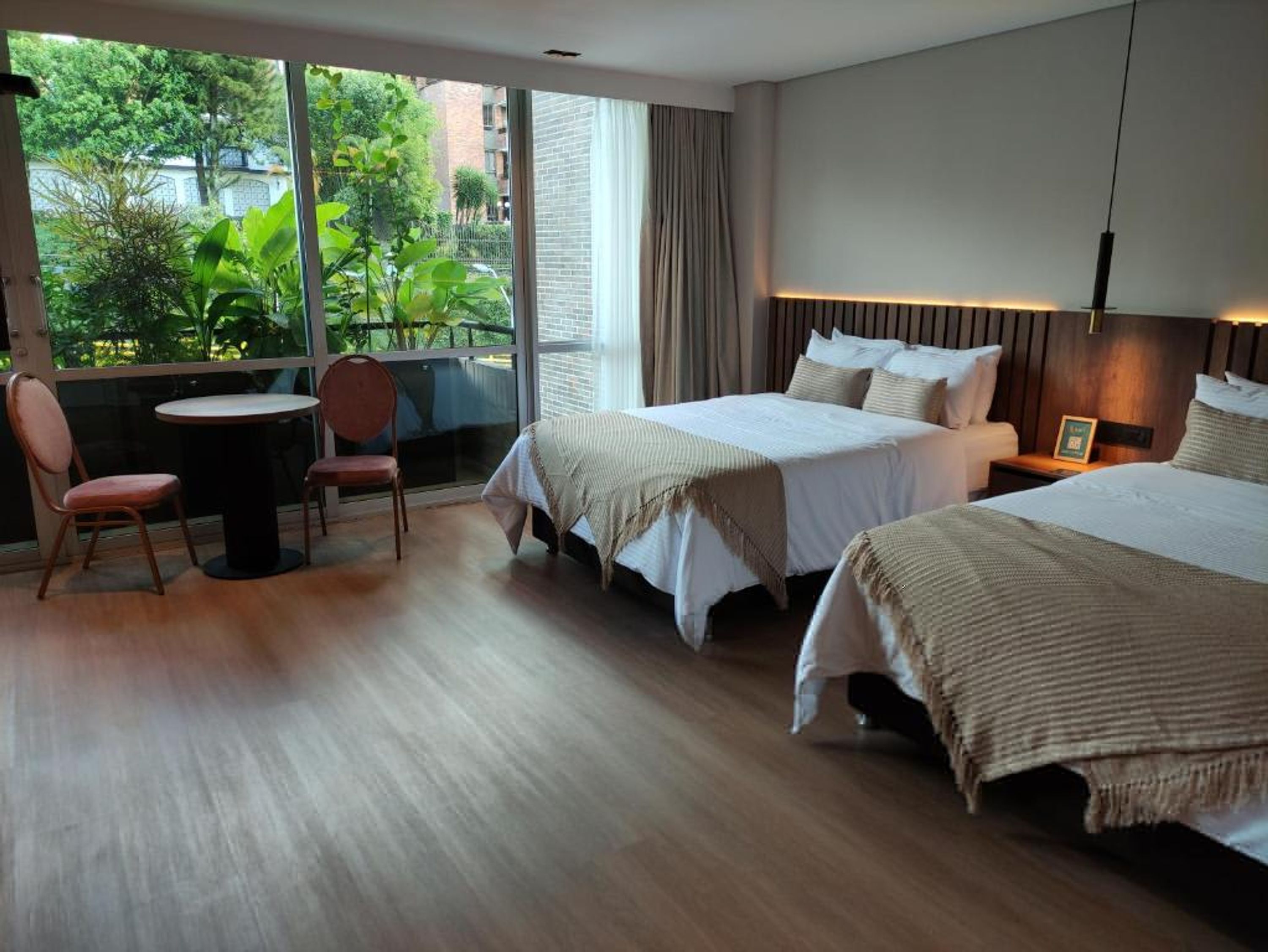 A hotel room with two beds, a small table, and a chair. There is a large window providing a view of greenery outside.
