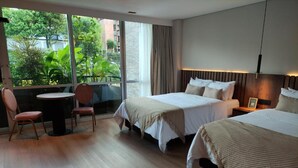 Une chambre d’hôtel avec deux lits, une petite table et une chaise. Une grande fenêtre offre une vue sur la verdure environnante.