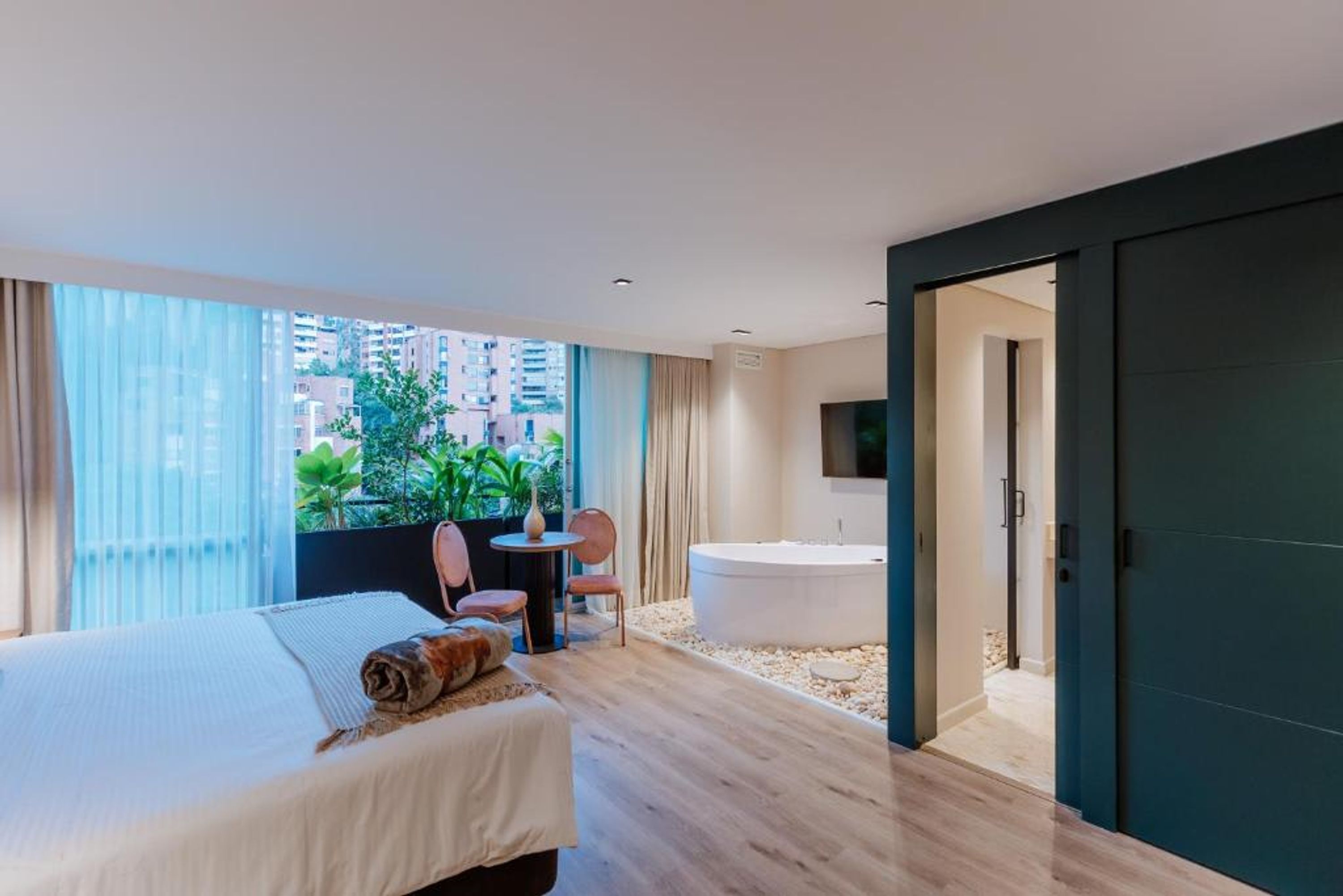 A modern hotel room with a large bed, a small table with chairs, a bathtub, and a view of the city through large windows.