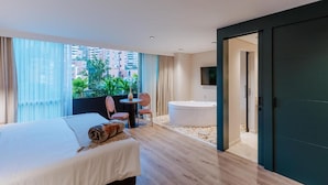 A modern hotel room with a large bed, a small table with chairs, a bathtub, and a view of the city through large windows. - w Loft Superior Mountain (Medellín)