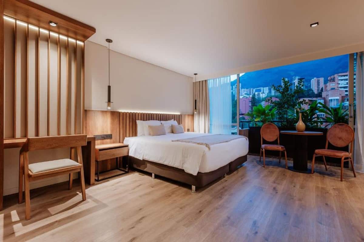 A modern bedroom with a large bed, wooden furniture, a desk, and a view of the city at dusk.