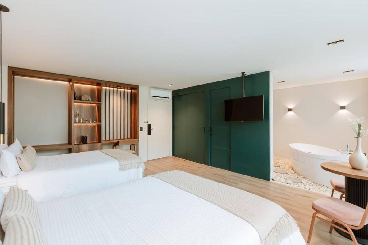 Ein modernes Hotelzimmer mit zwei Betten, einer Sitzecke mit Tisch und Stuhl, einer Badewanne und einem an der Wand montierten Fernseher.