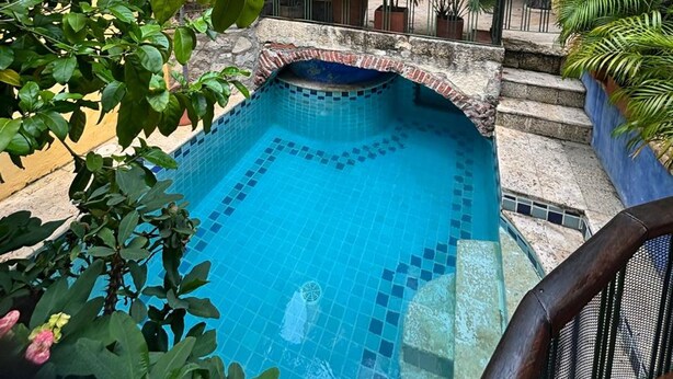 En gårdsplads med en swimmingpool, omgivet af frodig vegetation og en gul bygning med balkon.