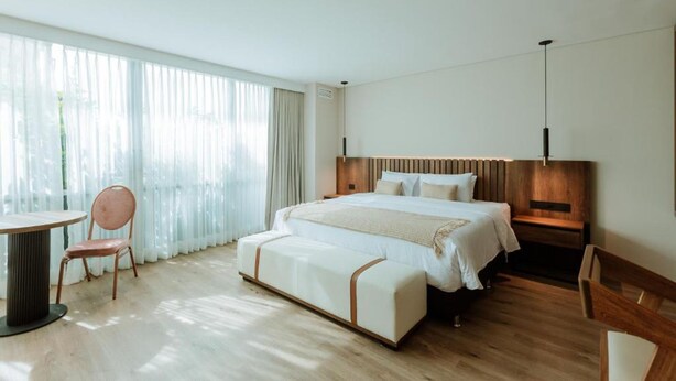 Una habitación de hotel moderna con una cama grande, una mesita de noche, una silla y vistas al exterior a través de cortinas translúcidas.