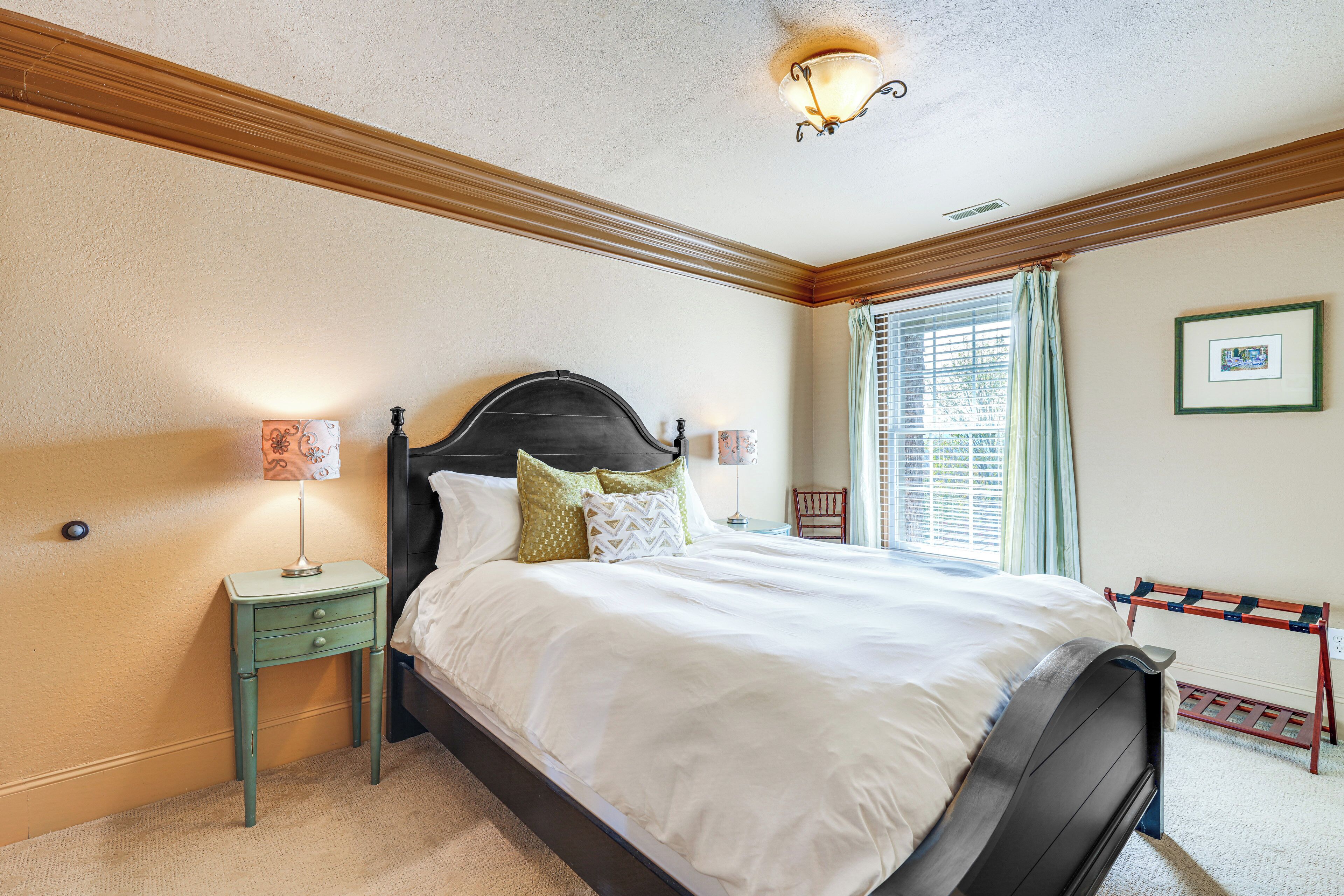 A bedroom with a large bed, two bedside tables, a window with blinds, and a wall-mounted picture.