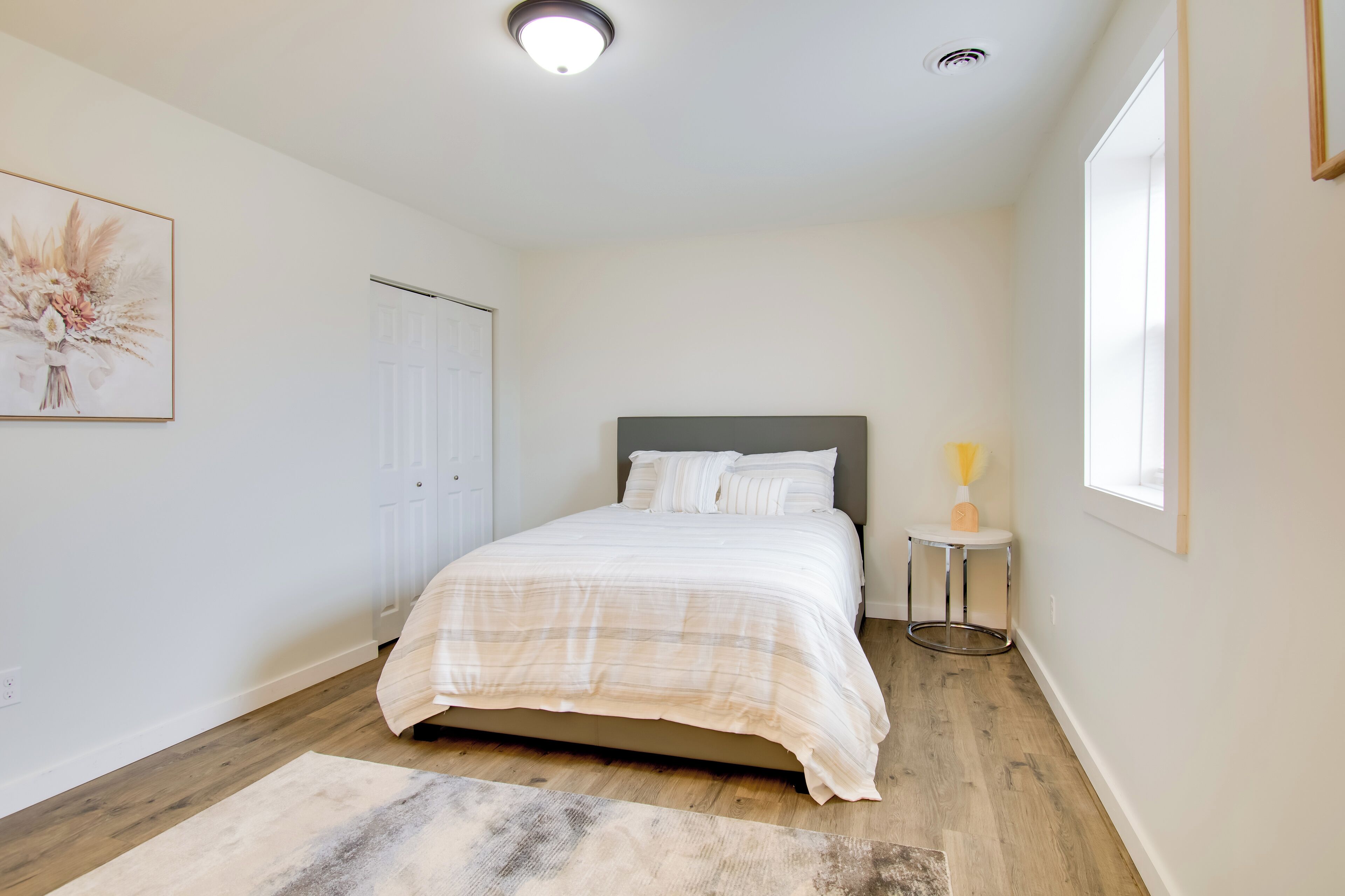 Quarto com cama, mesa de cabeceira com abajur, porta e quadro na parede.