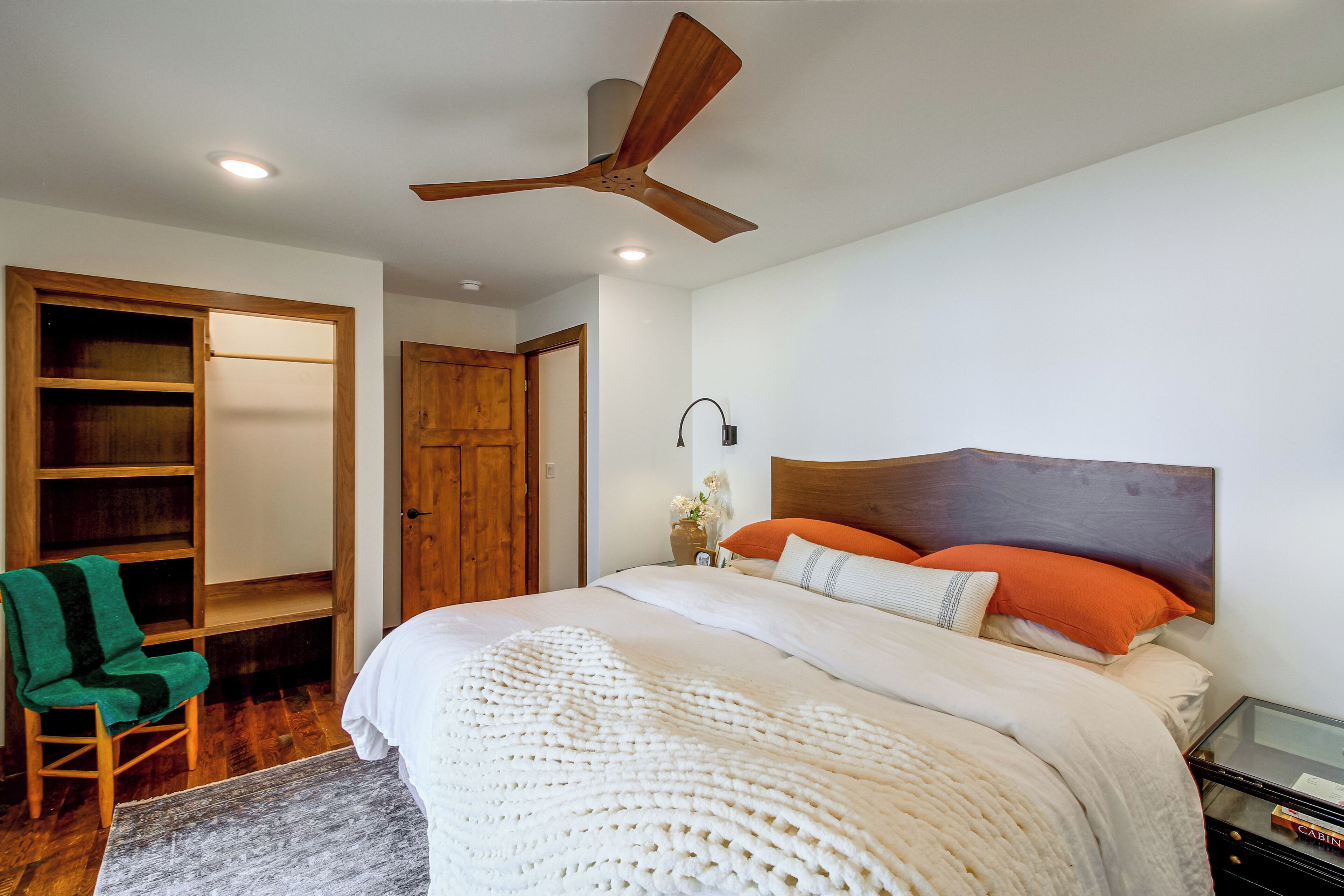 Une chambre à coucher comprenant un lit, une tête de lit en bois, une table de chevet, une chaise verte, une armoire en bois et un ventilateur de plafond.