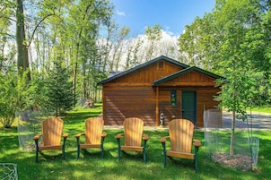 Una cabaña de madera con puerta verde, rodeada por una cerca de malla metálica, y tres sillas Adirondack de madera en el frente.
