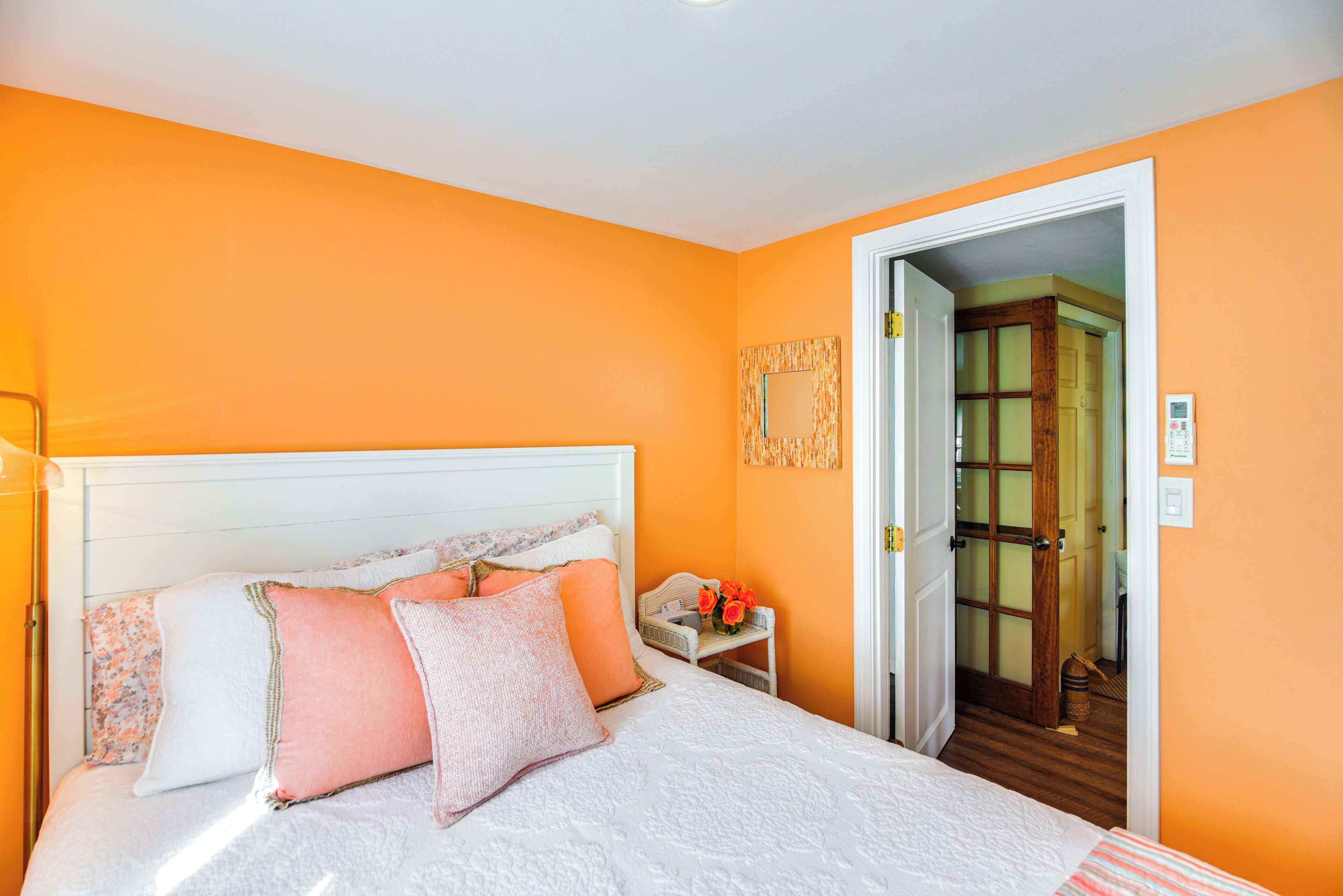 Un dormitorio con cama blanca, paredes anaranjadas y una puerta de madera que da a otra habitación.