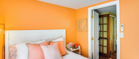 A bedroom with a white bed, orange walls, and a wooden door leading to another room.