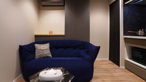 A modern living room with a dark blue sofa, a small table with plates, a wall-mounted shelf with a decorative item, and a built-in oven. - Real Life Iidabashi Stylish 1-bedroom 3 Mins Subway (Tokyo)