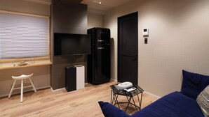 A modern living room with a flat-screen TV, a black refrigerator, a small table with a remote control, a blue sofa, and a white stool. - Real Life Iidabashi Stylish 1-bedroom 3 Mins Subway (Tokyo)