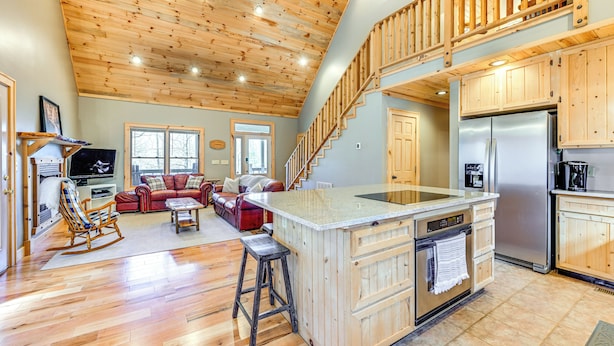 A spacious living area with a wooden ceiling, a kitchen with wooden cabinets, a large island, and a staircase leading to an upper level.