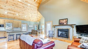 A spacious living area with a wooden ceiling, a fireplace, a television, and a kitchen in the background.