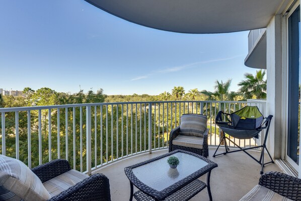 A balcony with wicker furniture, a glass table, and a view of palm trees and buildings. - Walk to White-sand Beach: Resort Condo in Destin! (Destin)