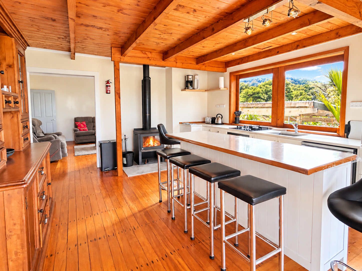 A modern kitchen with a central island, wooden flooring, and a large window offering a view of greenery.