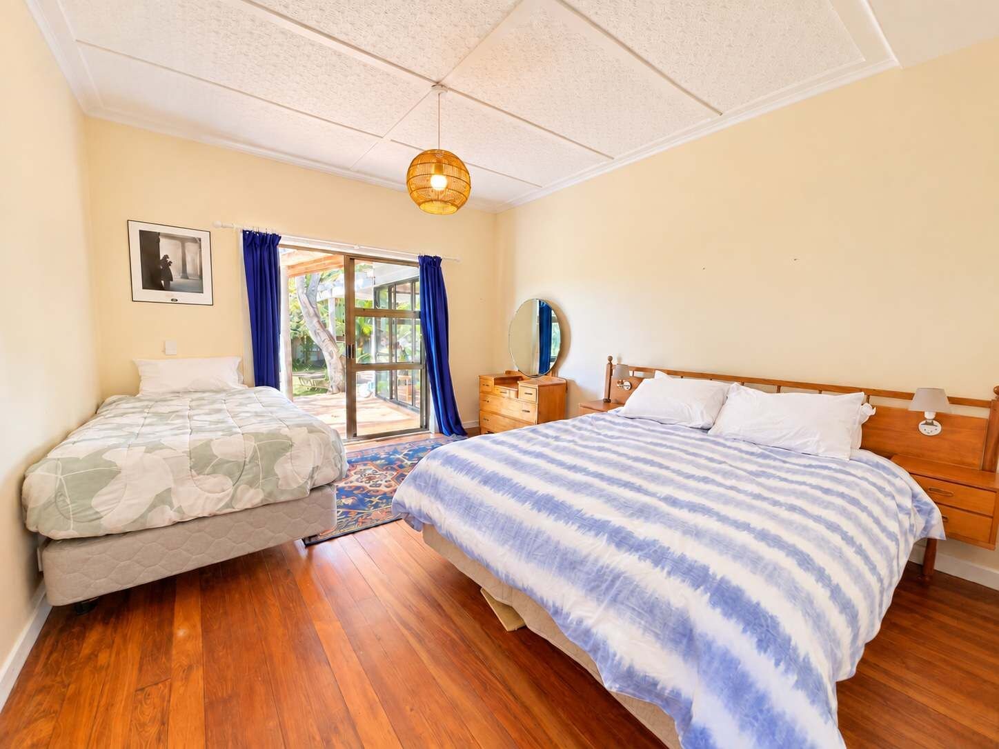 Une chambre avec deux lits, un plancher en bois, un luminaire au plafond, une fenêtre avec des rideaux et une porte donnant sur l’extérieur.