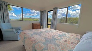 Ein Schlafzimmer mit Bett, Stuhl, Kommode und großen Fenstern mit Blick ins Grüne.