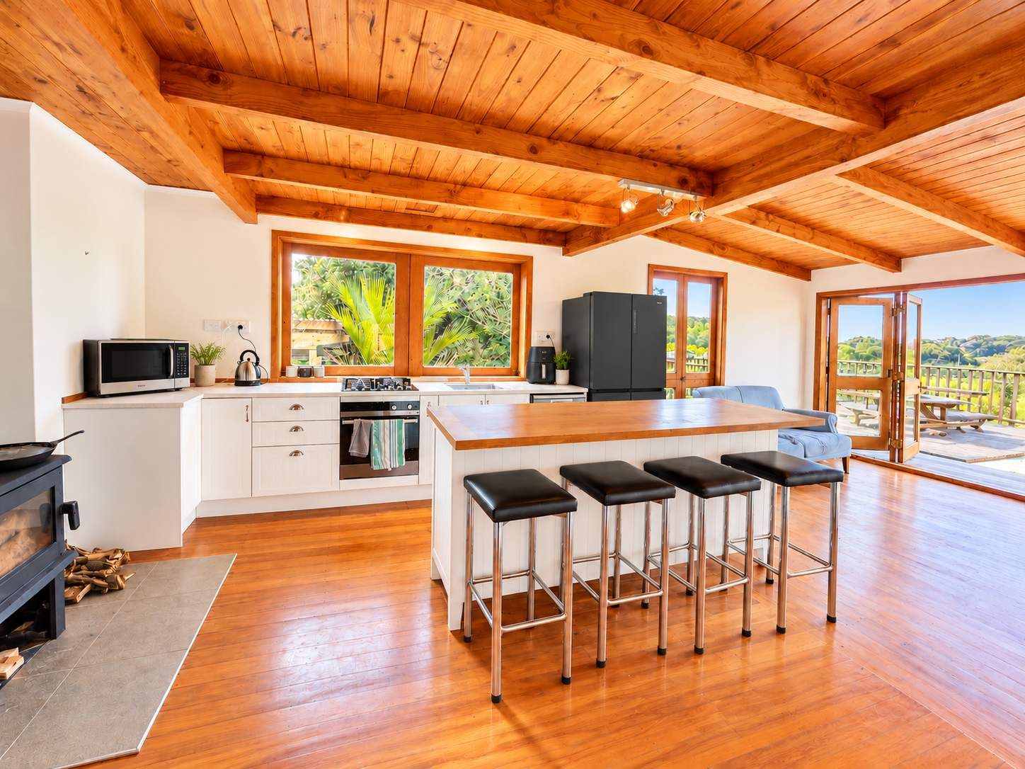 A modern kitchen with wooden beams, a central island with bar stools, a black refrigerator, and a view through large windows.
