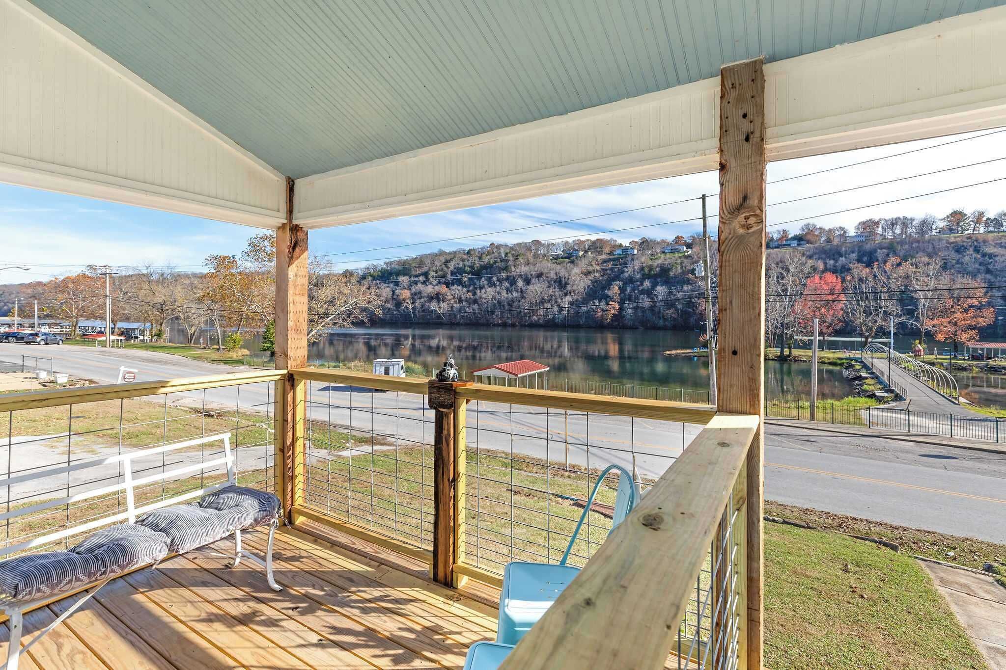 A covered deck with a bench, railing, and a view of a lake and road.