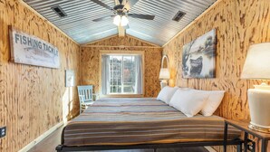 Una habitación con paredes de madera, una cama con ropa de cama a rayas, un ventilador de techo, una ventana y un cuadro.