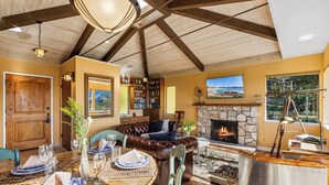 A dining area with a wooden table set for four, a leather sofa, a stone fireplace, and a bookshelf filled with books. - 3959 Hedgehog - Walk to Town and Beach, Ocean View (Carmel)