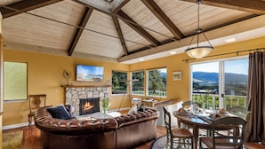A cozy living room with a stone fireplace, leather sofa, dining table, and large windows offering a view of the outdoors. - 3959 Hedgehog - Walk to Town and Beach, Ocean View (Carmel)
