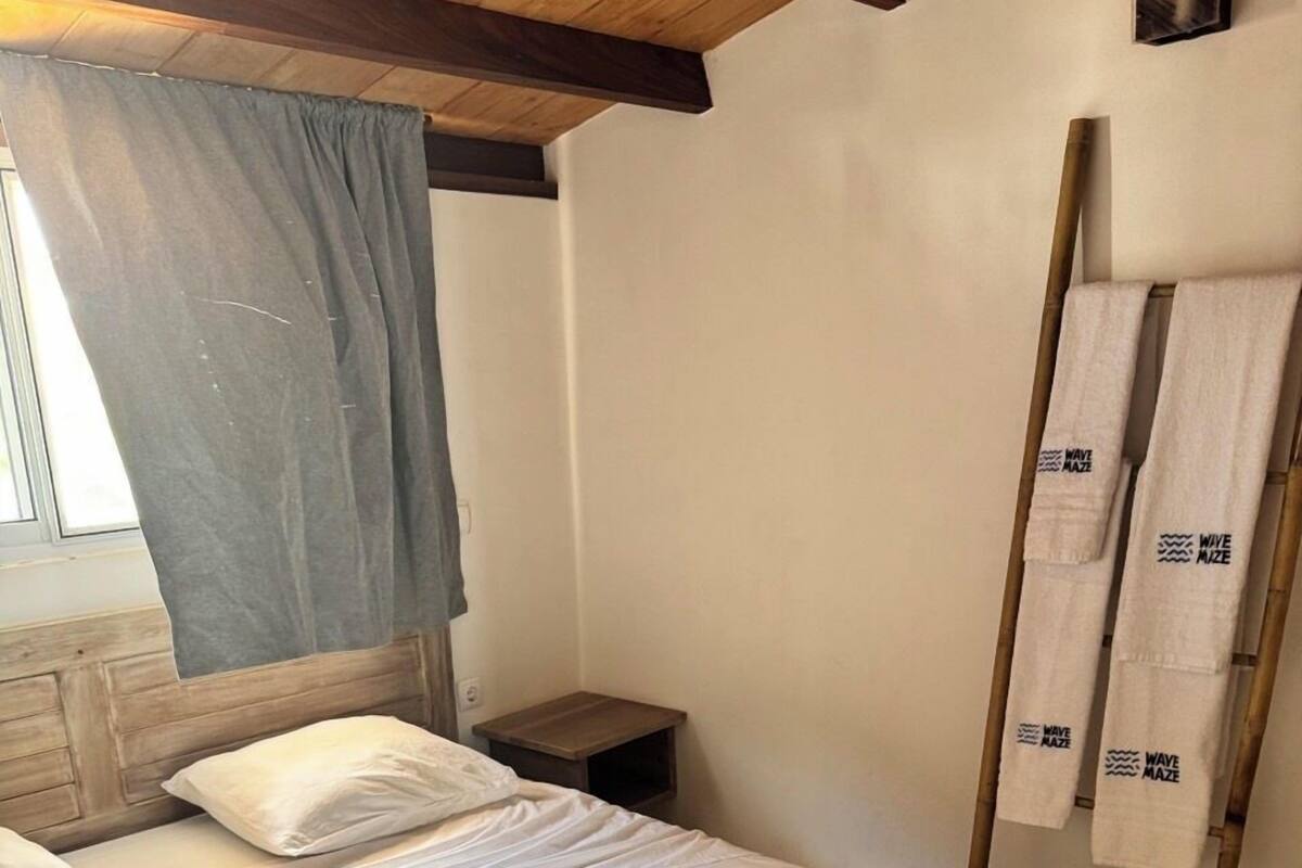 A bedroom with a bed, pillows, a nightstand, a window with curtains, and a towel rack with towels.