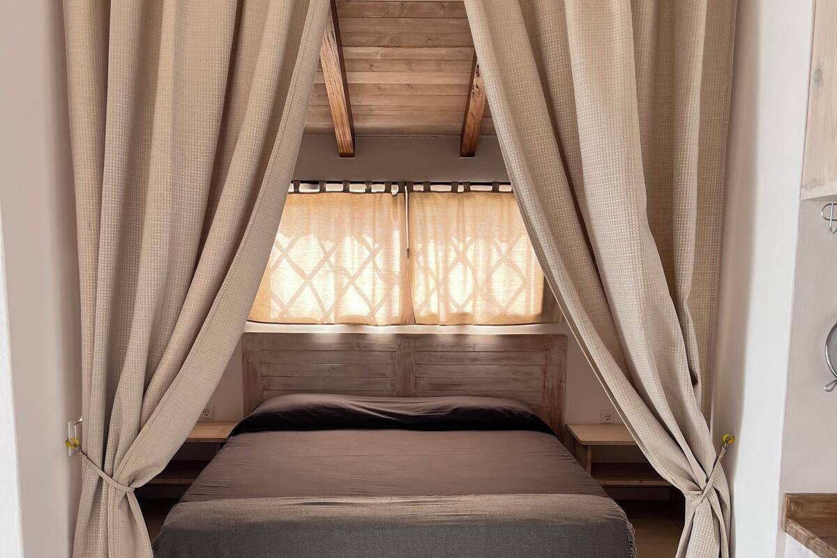 A cozy room with a bed, wooden ceiling beams, and beige curtains.