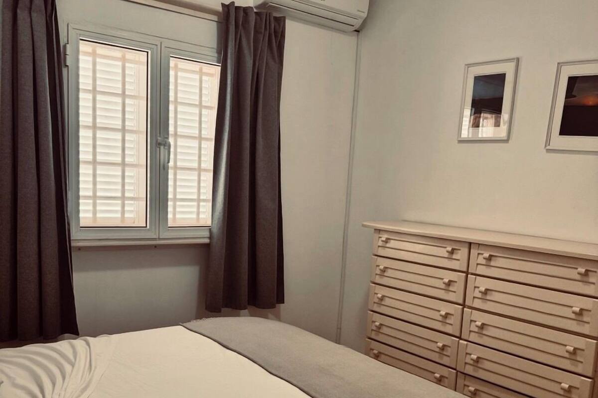 A bedroom with a bed, a dresser, an air conditioning unit, a window with curtains, and two framed pictures on the wall.