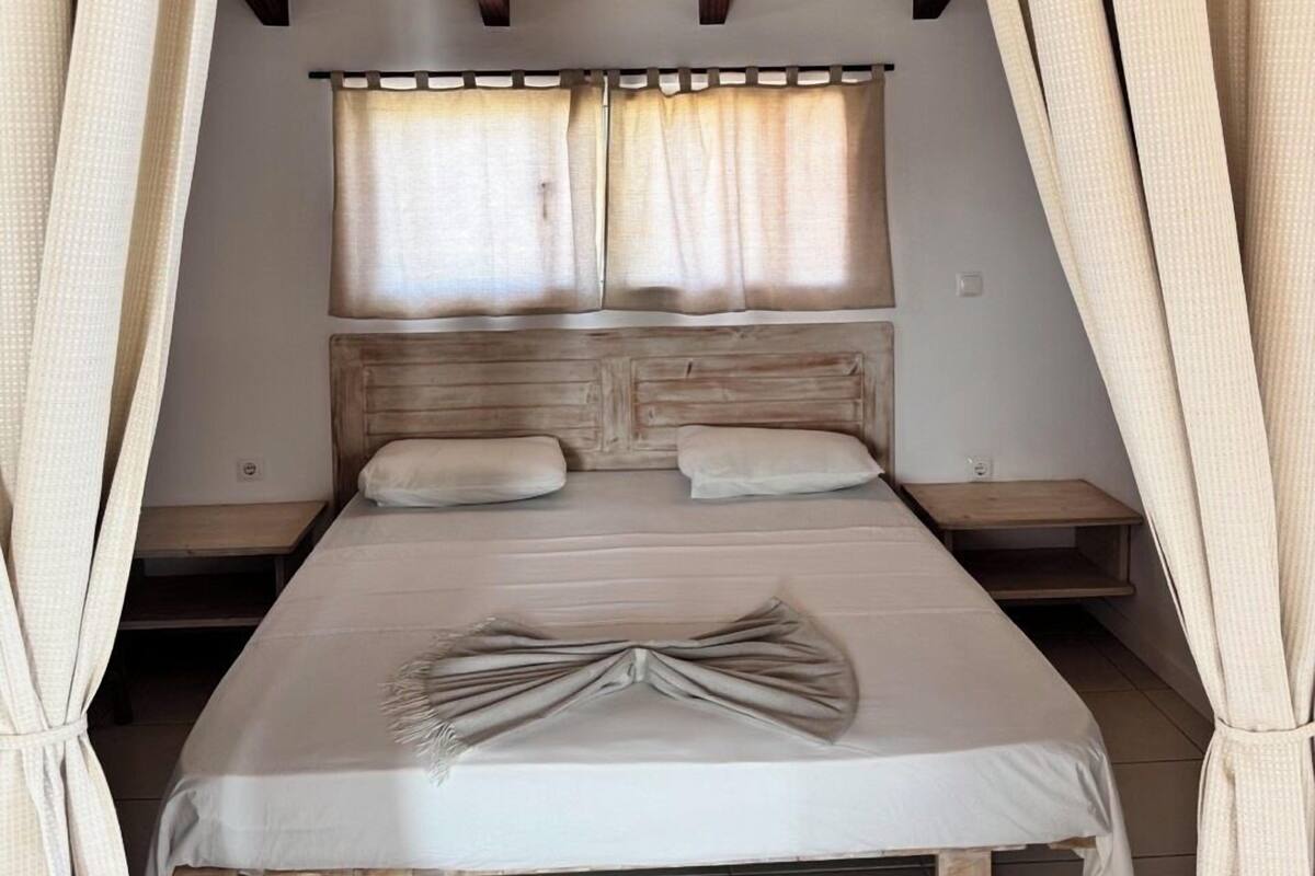 A bedroom with a wooden ceiling, a bed with a white bedspread, two bedside tables, and a window with curtains.