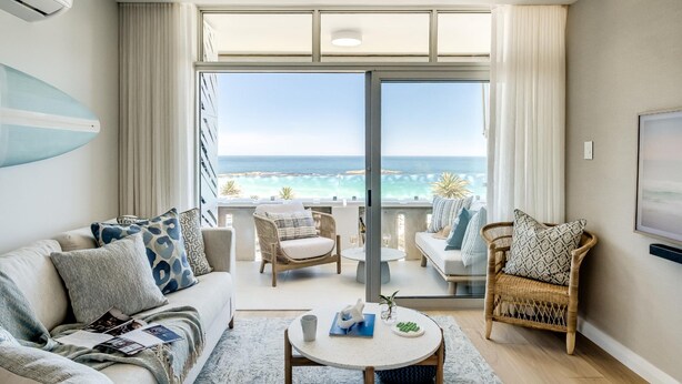 A modern living room with a view of the ocean, featuring a sofa, chairs, a coffee table, and a surfboard. - Apartment w Sea Views by Camps Bay Beach Kyanos (Cape Town)
