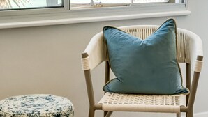 A room with a view of the ocean through a large window, a wicker chair with a cushion, and a round ottoman. - Apartment w Sea Views by Camps Bay Beach Kyanos (Cape Town)