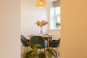 Un coin repas comprenant une table ronde, quatre chaises, un vase avec des fleurs séchées et un luminaire suspendu.