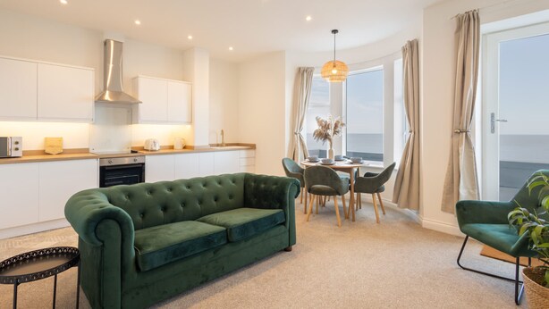 Ein modernes Wohnzimmer mit einem grünen Sofa, Essbereich, Küche und großen Fenstern.