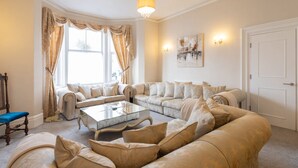 A spacious living room with a large sectional sofa, a glass coffee table, and a chandelier. There are two windows with curtains, a door, and a wall-mounted artwork. - Host Stay The Keep (Llandudno)