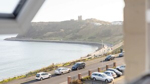 Une route côtière bordée de voitures garées, une plage et une colline surmontée d’une tour.