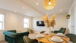 Un espace repas moderne avec une table ronde en bois, dressée pour quatre personnes, entourée de chaises à revêtement vert. Un luminaire tressé est suspendu au-dessus, et un téléviseur à écran plat est fixé au mur.