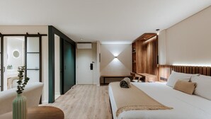 A modern hotel room with a large bed, a wooden headboard, a bedside table with a vase, and a seating area with a bench and chairs.
