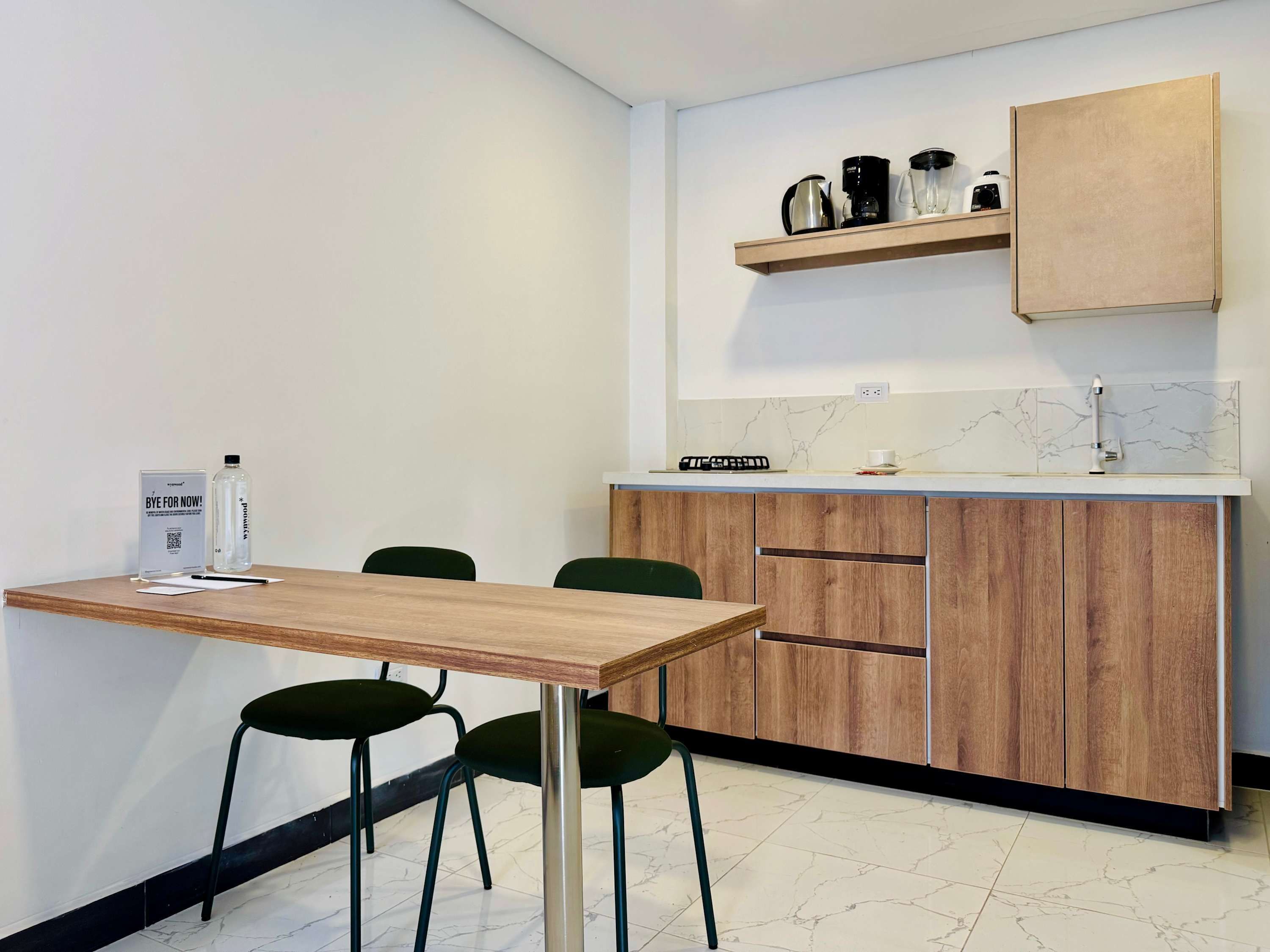 Una cocina moderna con una mesa de madera, dos sillas verdes, una cafetera y un estante con utensilios de cocina.