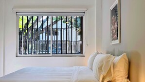 Quarto de hotel com uma cama, abajur e janela com vista para um edifício e árvores.