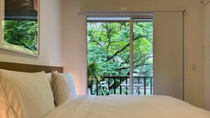 Ein Schlafzimmer mit einem großen Bett, Blick auf Bäume durch eine Schiebetür und ein kleiner Balkon mit Tisch und Stühlen.