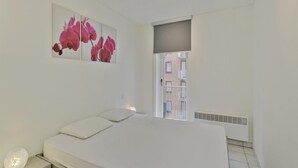 A bedroom with a bed, a nightstand, a wall-mounted light, and a window with a view of buildings. - Apartment in Blankenberge With Sea Views (Blankenberge)